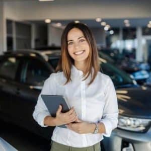 fusion sales sqr - Fusion Business College Smiling woman in a white shirt standing in a car dealership, holding a tablet in front of cars anchored in the showroom.