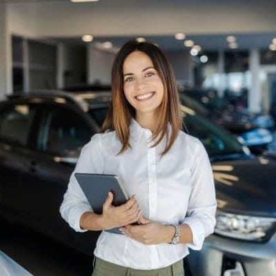 Smiling woman in a white shirt standing in a car dealership, holding a tablet in front of cars anchored in the showroom.