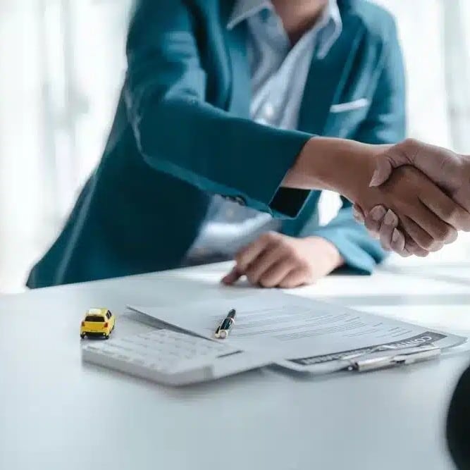 WA-Sales-License-sq - Fusion Business College Two people shake hands over a signed contract on a white desk, with a yellow toy car model and a pen nearby.